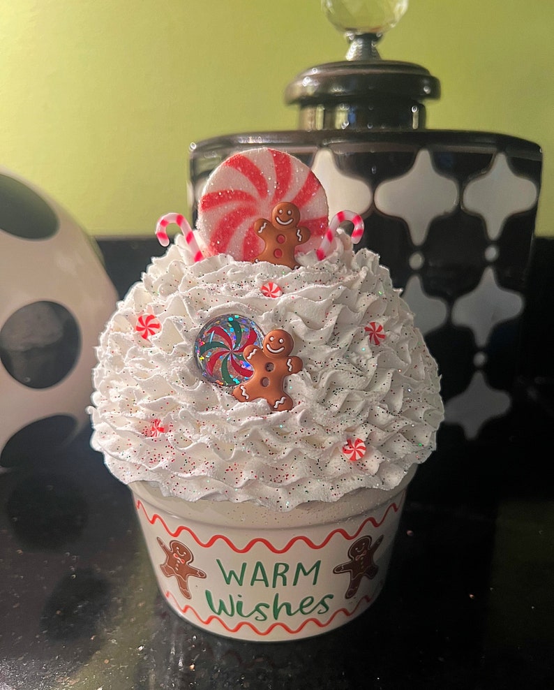 May include: A white ceramic cupcake container with a white frosting-like texture and red and white candy cane accents. The container has a red and white peppermint candy on top and two gingerbread men. The container has the words "Warm Wishes" written on the front.