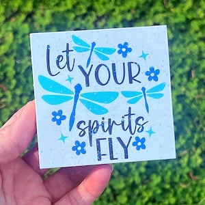 May include: Square white sign with the text "Let YOUR spirits FLY" in dark blue script. The sign features blue dragonfly and flower illustrations. The background has a speckled, glittery effect.