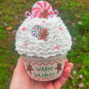 May include: White ceramic cup with a white and red candy cane, red and white peppermint candy, and gingerbread men decorations. The cup has a green and white wavy line design and the words "Warm Wishes" in green.