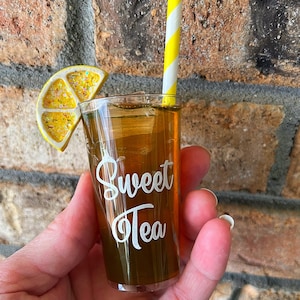 May include: A clear glass filled with iced tea and a lemon wedge, with a yellow and white striped straw. The glass has the words "Sweet Tea" written on it.