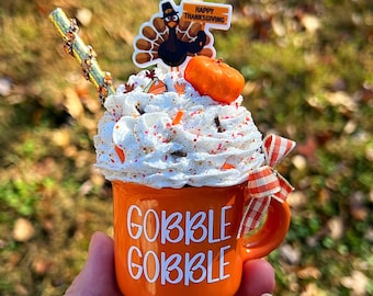 May include: Orange ceramic mug with white lettering that says "Gobble Gobble". The mug is topped with whipped cream, sprinkles, and a small orange pumpkin. A turkey figurine with a "Happy Thanksgiving" sign is on top of the whipped cream.