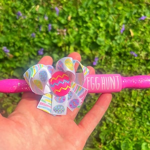 May include: Pink rolling pin with glitter and the words "Egg Hunt" written on it. A white and pastel pink bow with a pink and yellow Easter egg charm is attached to the rolling pin.