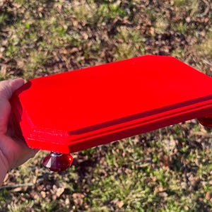 May include: A vibrant red, rectangular wooden tray with faceted, ruby-colored feet. The tray is held against a blurred background of green grass and brown leaves, showcasing its bright color and simple design.