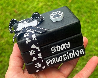 May include: Black wooden block set with a white paw print on top. The blocks are stacked and tied with a black and white ribbon with white paw prints and bone designs. The blocks read "Stay Pawsitive".