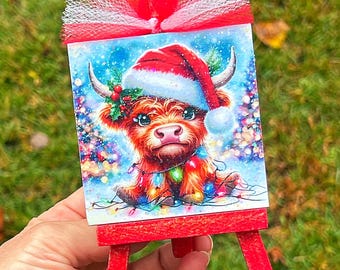 May include: A miniature red easel holds a Christmas-themed artwork. The artwork depicts a Highland cow wearing a Santa hat and Christmas lights. A red tulle bow adorns the top of the easel. The background is a blurred winter scene.