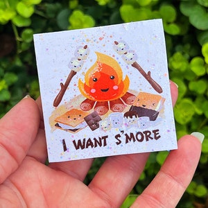 May include: A white square sticker with a cartoon illustration of a campfire with marshmallows and chocolate bars. The text "I WANT S'MORE" is printed on the sticker.