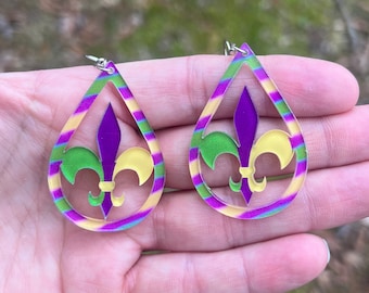 May include: A pair of teardrop-shaped earrings featuring a purple, green and yellow fleur-de-lis design. The earrings are made of clear acrylic and have a silver hook.