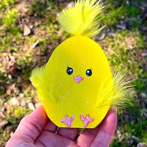 May include: A yellow wooden chick decoration with a fluffy feather top and side accents. The chick has black eyes, a pink beak, and pink feet. The decoration is speckled with glitter.