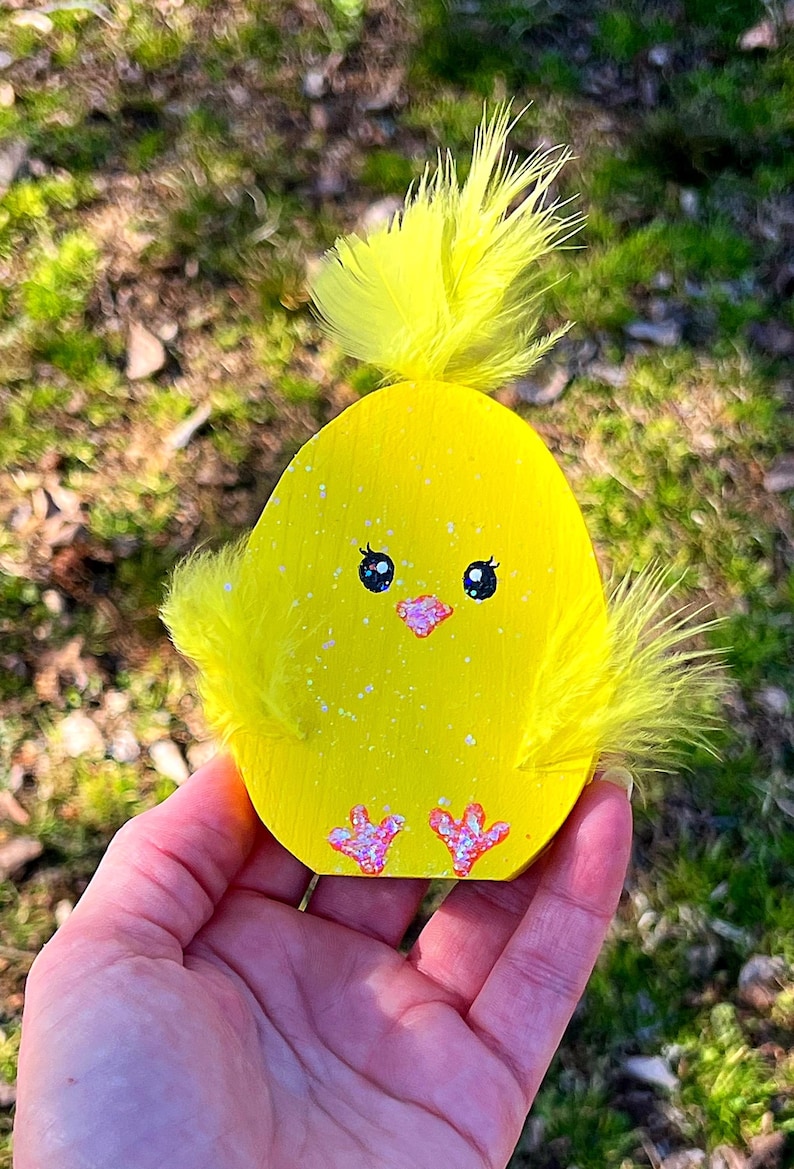 May include: A yellow wooden chick decoration with a fluffy feather top and side accents. The chick has black eyes, a pink beak, and pink feet. The decoration is speckled with glitter.