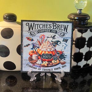 May include: A black and white sign with the text "Witches Brew Coffee Co." and a cartoon illustration of a pumpkin spice latte with Halloween decorations. The text at the bottom of the sign reads "Stirring up trouble since 1801."
