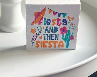 May include: White wooden sign with glitter and colorful text that reads "Fiesta and then Siesta". The sign features a sombrero, flowers, and a cactus.