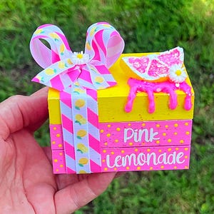 May include: A stack of three decorative wooden blocks painted yellow and pink with yellow polka dots. The top block features a pink grapefruit slice and white flower. A pink and white striped ribbon with lemon prints and a white flower bow.