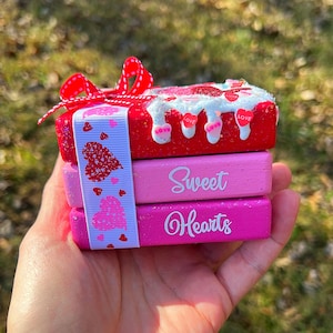 May include: Three stacked wooden boxes decorated with pink, red, and white glitter. The top box has a white icing design with red hearts and the words "LOVE" on it. The middle box is pink and says "Sweet" in white. The bottom box is pink and says "Hearts" in white.