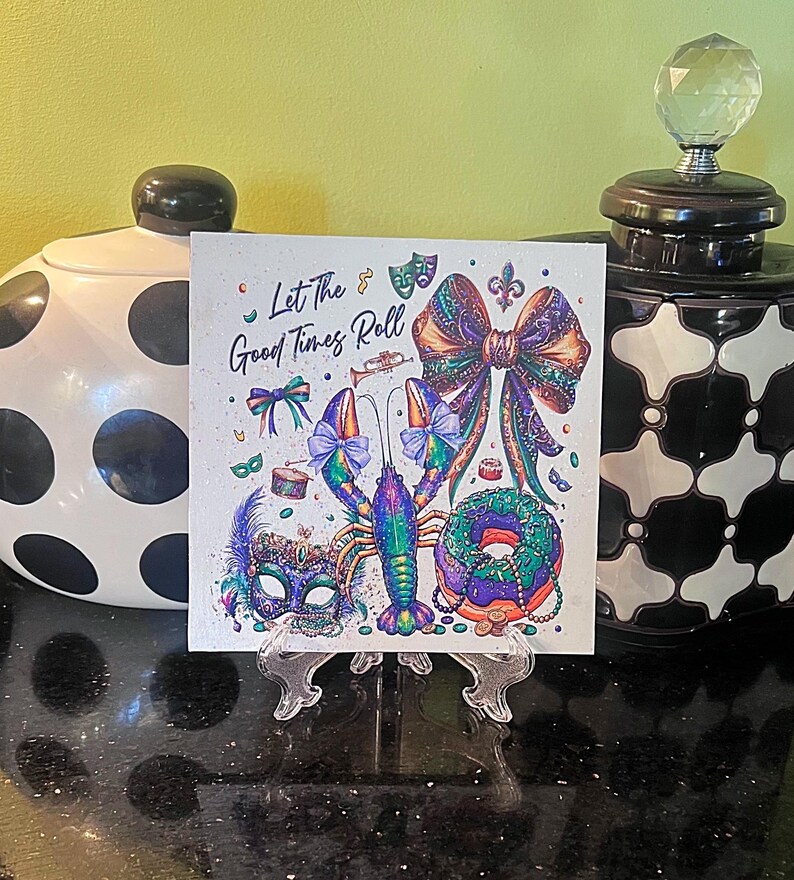 Pode incluir: Impress&atilde;o art&iacute;stica quadrada com tema Mardi Gras. A impress&atilde;o apresenta uma ilustra&ccedil;&atilde;o colorida de uma lagosta, la&ccedil;o, m&aacute;scara e donut. O texto diz "Let The Good Times Roll". A impress&atilde;o &eacute; exibida em um suporte transparente.
