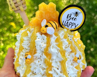 May include: A white whipped cream cake decorated with yellow honey, gold glitter, and small plastic bees. The cake is topped with a honeycomb, a wooden honey dipper, and a round black and white sign that says "Bee Happy".
