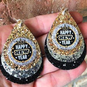 May include: A pair of layered teardrop earrings with a New Year's theme. The earrings feature a black, silver, and gold glitter design. The center circle has the words "Happy New Year" in a black and gold design.
