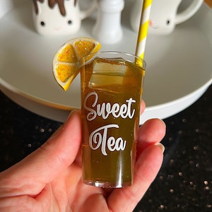 May include: A miniature glass of iced tea with a lemon wedge and a yellow and white striped straw. The glass is labeled "Sweet Tea".