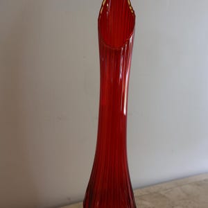 May include: A tall, slender, ruby red glass vase featuring a vertical ribbed texture throughout its body. The vase has a flared, scalloped rim at the top and a rounded, bulbous base that tapers into a long, narrow neck, crafted from translucent glass.