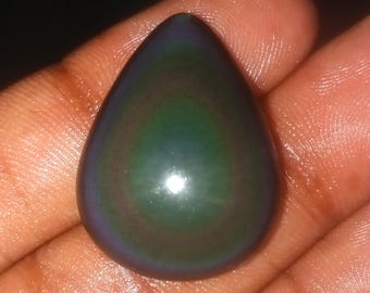 May include: A teardrop-shaped, polished gemstone with a dark, iridescent sheen. The stone displays a blend of deep blues, greens, and purples, with a glossy surface. The gemstone is held in a hand, showcasing its size and shape.