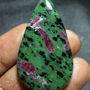 May include: A polished, teardrop-shaped gemstone featuring a vibrant green base speckled with black inclusions and striking streaks of deep red. The stone's surface is smooth and reflective, showcasing a natural pattern. This gemstone is ideal for jewelry making.