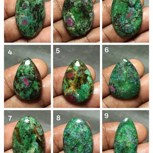 May include: Nine polished ruby zoisite gemstones in various shapes. The stones display a mix of green, red, and black colors. The gemstones are oval, teardrop, and elongated shapes. The stones are held in a hand.