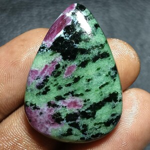 Pode incluir: Uma pedra preciosa polida em forma de gota, apresentando uma mistura de cores. A pedra tem uma base verde com inclusões pretas e rosa. A gema é segurada em uma mão, mostrando sua beleza natural e seu padrão único.
