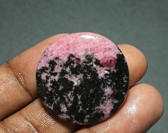 May include: A polished, round gemstone with a striking color contrast. The top half is a vibrant pink, transitioning to a speckled black and pink lower half. The stone is held in a hand, showcasing its smooth, reflective surface.