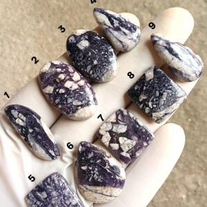 Puede incluir: Una colección de nueve piedras preciosas pulidas de varias formas, sostenidas en una mano enguantada. Las piedras presentan una mezcla de patrones morados y blancos. Las formas incluyen óvalo, lágrima y cuadrado, mostrando variaciones naturales.