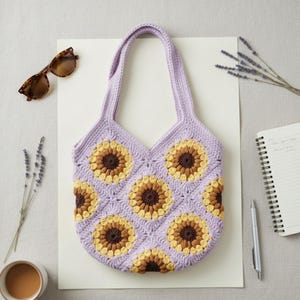 May include: A handmade crochet tote bag in a light purple color, featuring a sunflower pattern in yellow and brown. The bag has two handles and is sitting on a white surface. Sunglasses and a notebook are also visible.