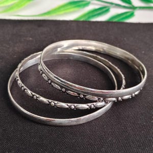 May include: A set of three silver-toned metal bangles. One bangle features a smooth, plain surface, while the other two bangles are decorated with a repeating pattern of small, raised oval shapes and tiny spheres along the entire outer circumference.