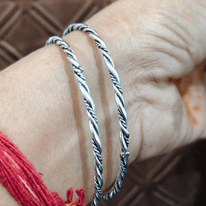 Könnte beinhalten: Zwei silberfarbene Armreifen mit einem gedrehten, seilartigen Design. Die Armbänder haben eine polierte metallische Oberfläche mit abgedunkelten vertieften Bereichen zwischen den Drehungen, was den runden Metallbändern eine Textur verleiht.