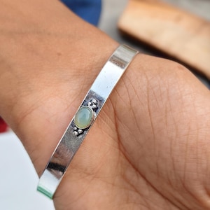 Könnte beinhalten: Ein silberfarbenes Armreif-Armband mit einem ovalen, hellgrünen Edelstein in der Mitte. Der Edelstein ist von einer dekorativen Fassung mit kleinen, runden Verzierungen umgeben. Das Armband wird am Handgelenk getragen.