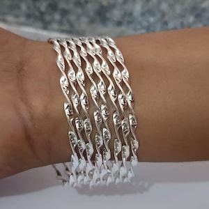Peut inclure: Ensemble de bracelets argentés avec un motif de corde torsadée. Les bracelets sont portés sur un poignet, mettant en valeur leur aspect métallique réfléchissant. Le design présente une série de motifs ondulés répétés, créant un aspect texturé.