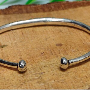 K&ouml;nnte beinhalten: Ein silberfarbenes Armband in offener Form mit glatter, polierter Oberfl&auml;che und abgerundeten, knollenartigen Enden. Das Metallband weist eine leicht unregelm&auml;&szlig;ige, organische Form auf, die sich zu einem Kreis biegt und eine reflektierende Oberfl&auml;che zeigt.