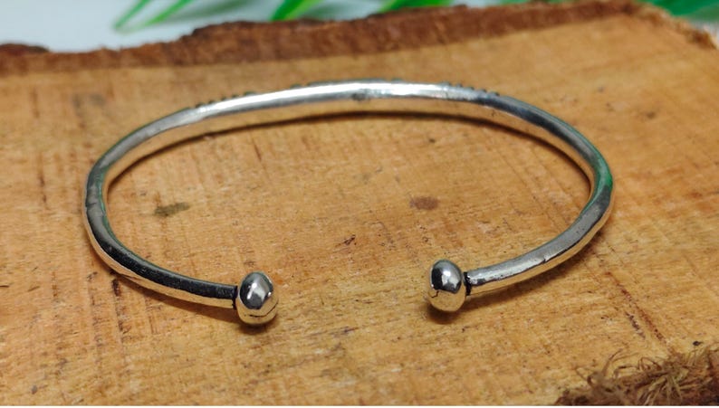 K&ouml;nnte beinhalten: Ein silberfarbenes Armband in offener Form mit glatter, polierter Oberfl&auml;che und abgerundeten, knollenartigen Enden. Das Metallband weist eine leicht unregelm&auml;&szlig;ige, organische Form auf, die sich zu einem Kreis biegt und eine reflektierende Oberfl&auml;che zeigt.