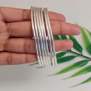 Könnte beinhalten: Ein Set aus sieben silbernen Armreifen, jeweils mit glatter, reflektierender Oberfläche. Die Armreifen sind gestapelt, was ihr schlichtes, elegantes Design hervorhebt. Der Schmuck wird vor einem weißen Hintergrund mit grünem Laub gezeigt.