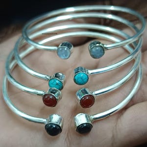 Könnte beinhalten: Eine Sammlung silberfarbener Metallarmreifen. Jeder Armreif hat an jedem Ende einen Edelstein, darunter blaue, türkisfarbene, rote und schwarze Steine. Die Armreifen werden auf einer hellbraunen Oberfläche präsentiert.