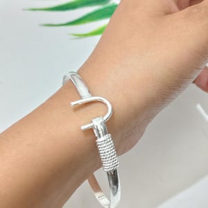 Könnte beinhalten: Ein silberfarbenes Armband mit Haken- und Ösenverschluss. Das Armband im Bangle-Stil hat ein glattes, abgerundetes Band. Der Verschluss hat ein strukturiertes, gewickeltes Design. Das Armband wird am Handgelenk getragen.