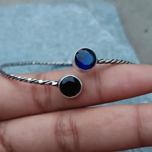 Könnte beinhalten: Ein silberfarbenes Armband mit gedrehtem Design. Es hat einen runden, dunkelblauen Edelstein und einen runden, schwarzen Edelstein. Das Armband wird auf der Hand einer Person gezeigt.