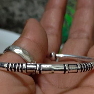K&ouml;nnte beinhalten: Ein silberfarbenes Armband in Manschettenform mit polierter Oberfl&auml;che und schwarzen, eingravierten linearen sowie rechteckigen Mustern entlang des Bandes. Das Armband hat abgerundete Enden und eine glatte, geschwungene Struktur.