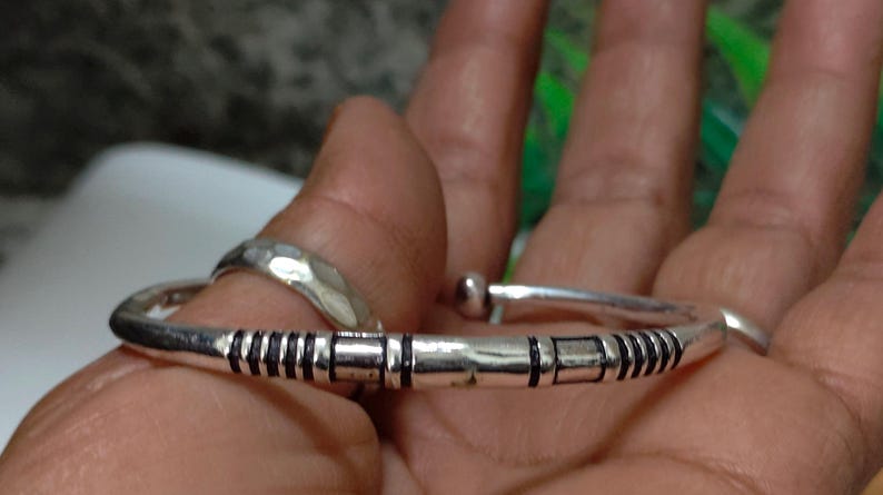 K&ouml;nnte beinhalten: Ein silberfarbenes Armband in Manschettenform mit polierter Oberfl&auml;che und schwarzen, eingravierten linearen sowie rechteckigen Mustern entlang des Bandes. Das Armband hat abgerundete Enden und eine glatte, geschwungene Struktur.