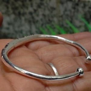 K&ouml;nnte beinhalten: Ein silberfarbenes, offenes Armband mit einem glatten, abgerundeten Reifen und kleinen kugelf&ouml;rmigen Endst&uuml;cken. Das Armband weist dezente, eingravierte lineare Details auf der Au&szlig;enfl&auml;che auf, die f&uuml;r ein schlichtes und poliertes Erscheinungsbild sorgen.