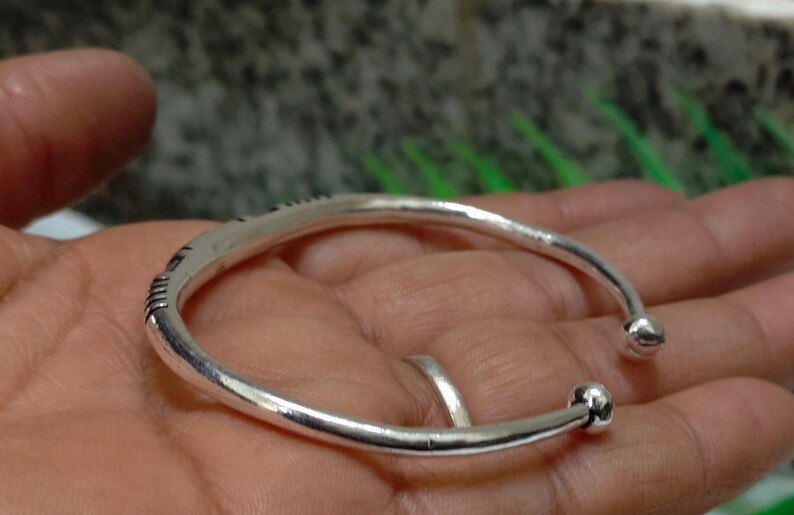 K&ouml;nnte beinhalten: Ein silberfarbenes, offenes Armband mit einem glatten, abgerundeten Reifen und kleinen kugelf&ouml;rmigen Endst&uuml;cken. Das Armband weist dezente, eingravierte lineare Details auf der Au&szlig;enfl&auml;che auf, die f&uuml;r ein schlichtes und poliertes Erscheinungsbild sorgen.
