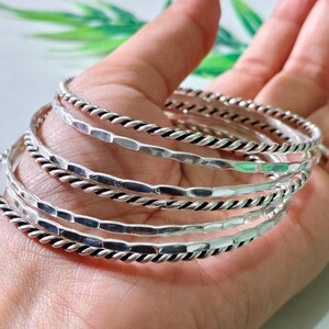 Könnte beinhalten: Ein Set aus fünf silberfarbenen Armreifen mit abwechselnden Designs. Drei Armreifen haben eine gedrehte Seilstruktur, während zwei Armreifen eine gehämmerte, facettierte Oberfläche aufweisen. Die Armreifen sind rund und haben ein poliertes Finish.