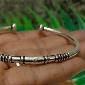 K&ouml;nnte beinhalten: Ein silberfarbenes, offenes Armband mit polierter Oberfl&auml;che und dunklen, vertieften linearen Details im Mittelteil. Das Armband hat abgerundete, glatte Enden an der &Ouml;ffnung und bildet so ein schlichtes, durchgehendes kreisf&ouml;rmiges Band.