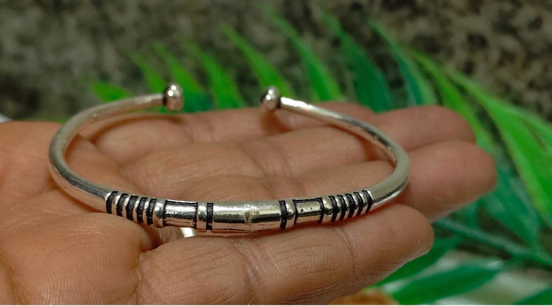 K&ouml;nnte beinhalten: Ein silberfarbenes, offenes Armband mit polierter Oberfl&auml;che und dunklen, vertieften linearen Details im Mittelteil. Das Armband hat abgerundete, glatte Enden an der &Ouml;ffnung und bildet so ein schlichtes, durchgehendes kreisf&ouml;rmiges Band.