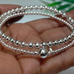 Könnte beinhalten: Ein silbernes Armband mit zwei Strängen aus kleinen, runden Perlen. Das Armband wird in einer Hand gehalten, wodurch seine glänzende Oberfläche hervorgehoben wird. Das Design umfasst eine größere Perle als Mittelpunkt.