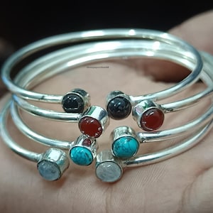 Könnte beinhalten: Eine Sammlung silberfarbener Armreifen, jeder mit einem anderen Edelstein verziert. Die Edelsteine umfassen schwarze, rote, türkisfarbene und hellblaue Steine. Die Armreifen sind gestapelt und auf einer Hand präsentiert.
