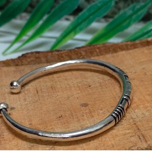 K&ouml;nnte beinhalten: Ein silberfarbenes, offenes Armband mit glatter, polierter Oberfl&auml;che und zwei kleinen kugelf&ouml;rmigen Endst&uuml;cken. Das Band ist an einer Seite mit einer Reihe von eingravierten, dunkel gef&auml;rbten parallelen Linien und geometrischen Mustern verziert.