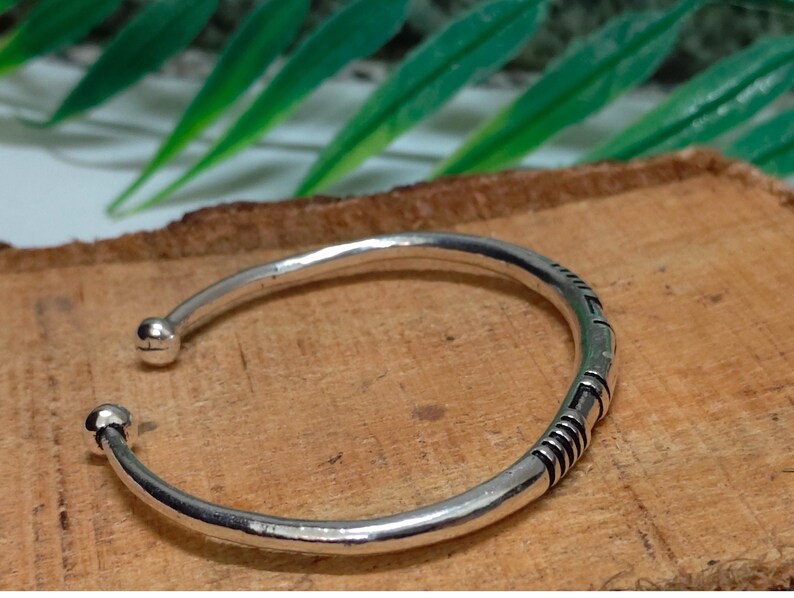 K&ouml;nnte beinhalten: Ein silberfarbenes, offenes Armband mit glatter, polierter Oberfl&auml;che und zwei kleinen kugelf&ouml;rmigen Endst&uuml;cken. Das Band ist an einer Seite mit einer Reihe von eingravierten, dunkel gef&auml;rbten parallelen Linien und geometrischen Mustern verziert.